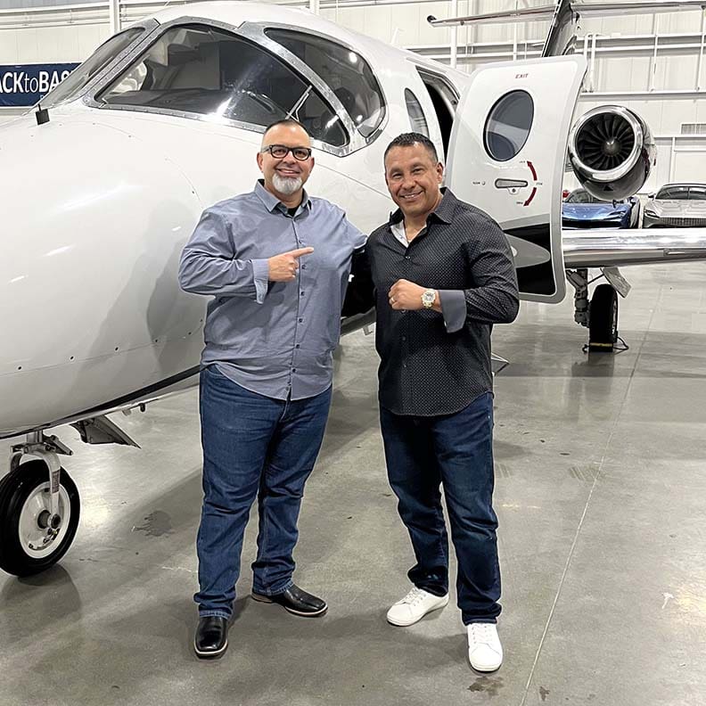 Two men are smiling and pointing at each other while standing in front of a sleek, white private jet inside a hangar. This candid "About Us" moment showcases their camaraderie, with both casually dressed in jeans and shirts. The aircraft's open door and polished floor gleam in the background.