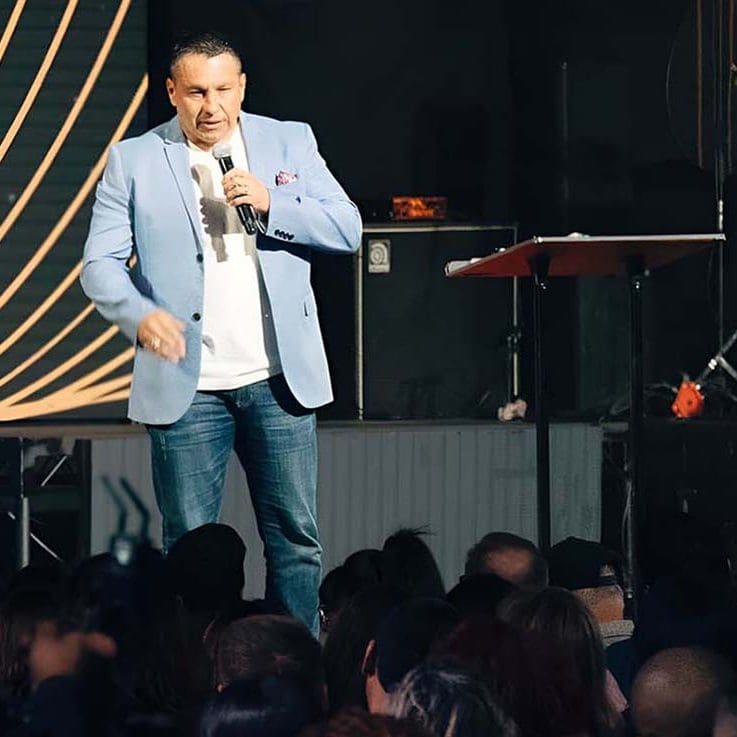 Daniel Gomez wearing a light blue blazer, white shirt, and blue jeans is speaking into a microphone on stage. He appears engaged with the audience, who are seated in front of him. With a music stand and some equipment in the background, the setting suggests an indoor coaching event. Daniel Gomez wearing a light blue blazer, white shirt, and blue jeans is speaking into a microphone on stage. He appears engaged with the audience, who are seated in front of him. With a music stand and some equipment in the background, the setting suggests an indoor coaching event.