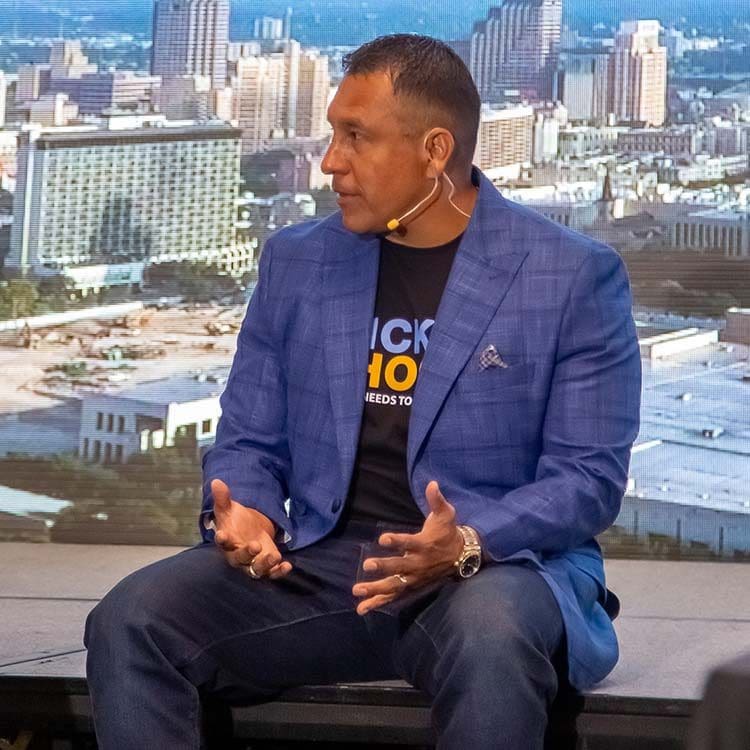 Daniel Gomez sits onstage, coaching the audience with animated hand gestures. He has a headset microphone, and a cityscape with tall buildings is visible in the background.